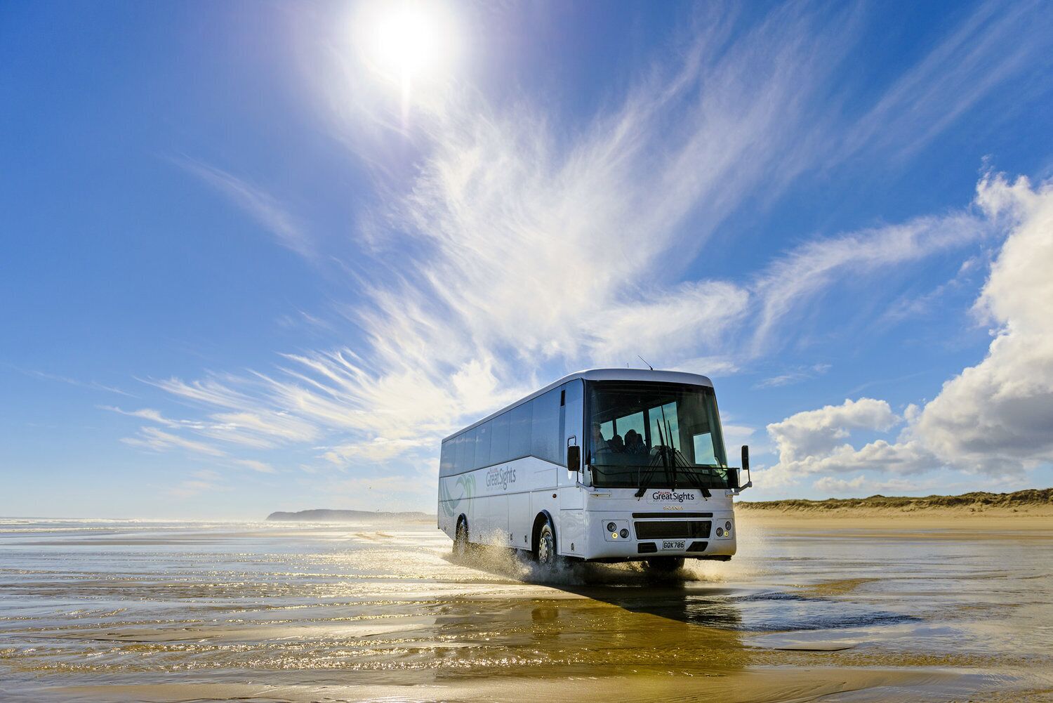 A white bus with "Great Sights" branding driving through shallow water on a beach.