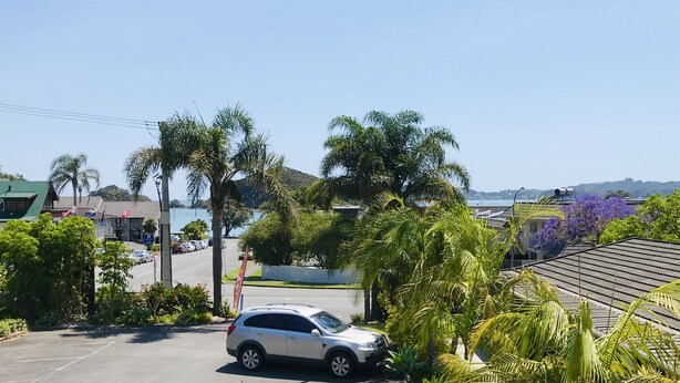 A parking lot with two cars, palm trees, and a view of a body of water and hills in the background. - Bounty Motel (Paihia)