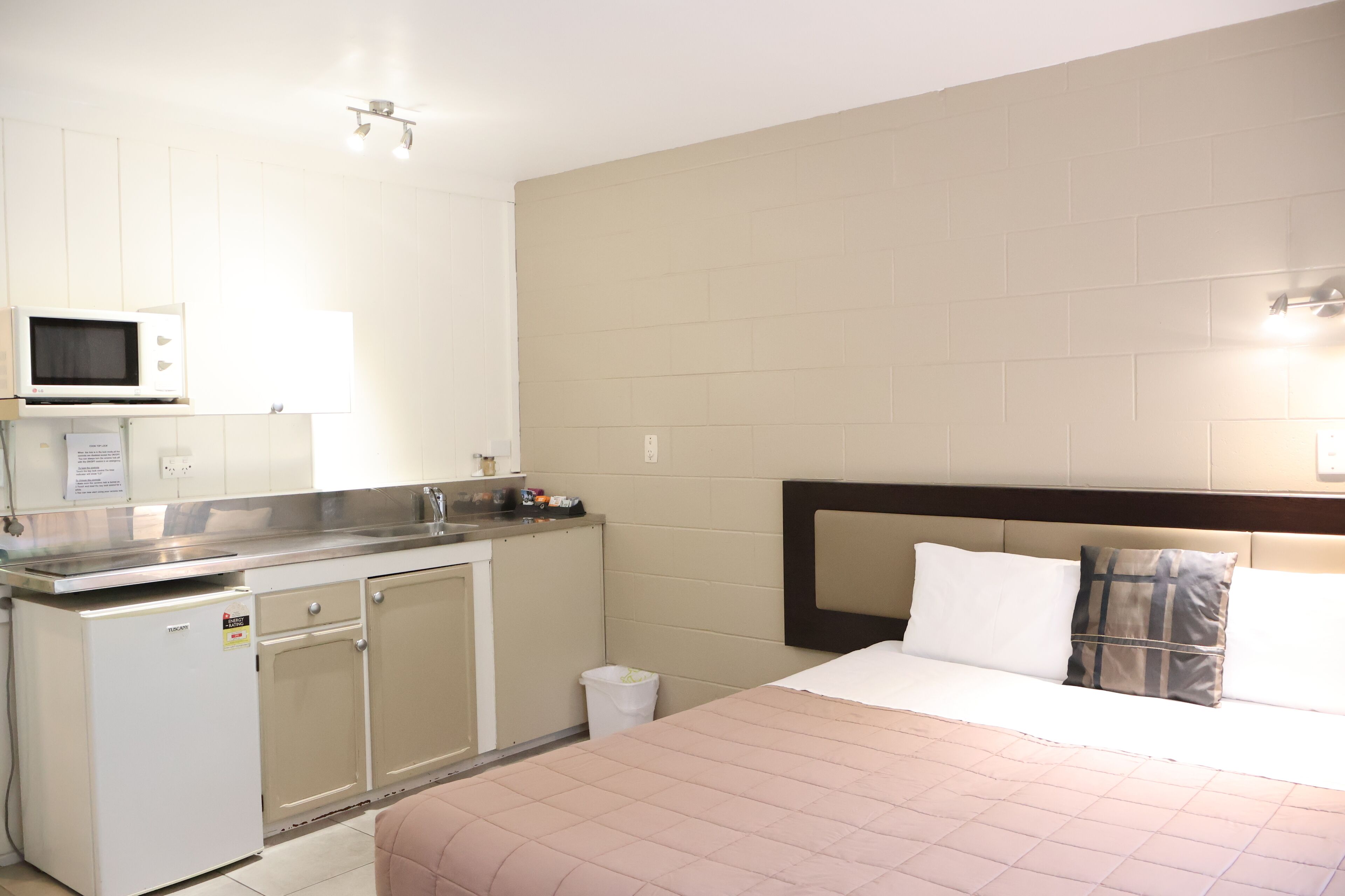 A hotel room with a kitchenette, a bed with two pillows, and a plain wall.