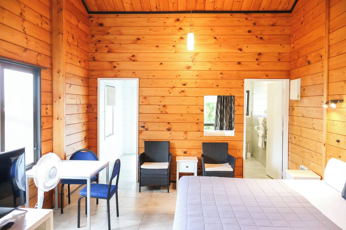 Ein Zimmer in einer Holzhütte mit einem Bett, einem Schreibtisch mit Stuhl, zwei Sesseln, einem kleinen Tisch und einem Fenster.