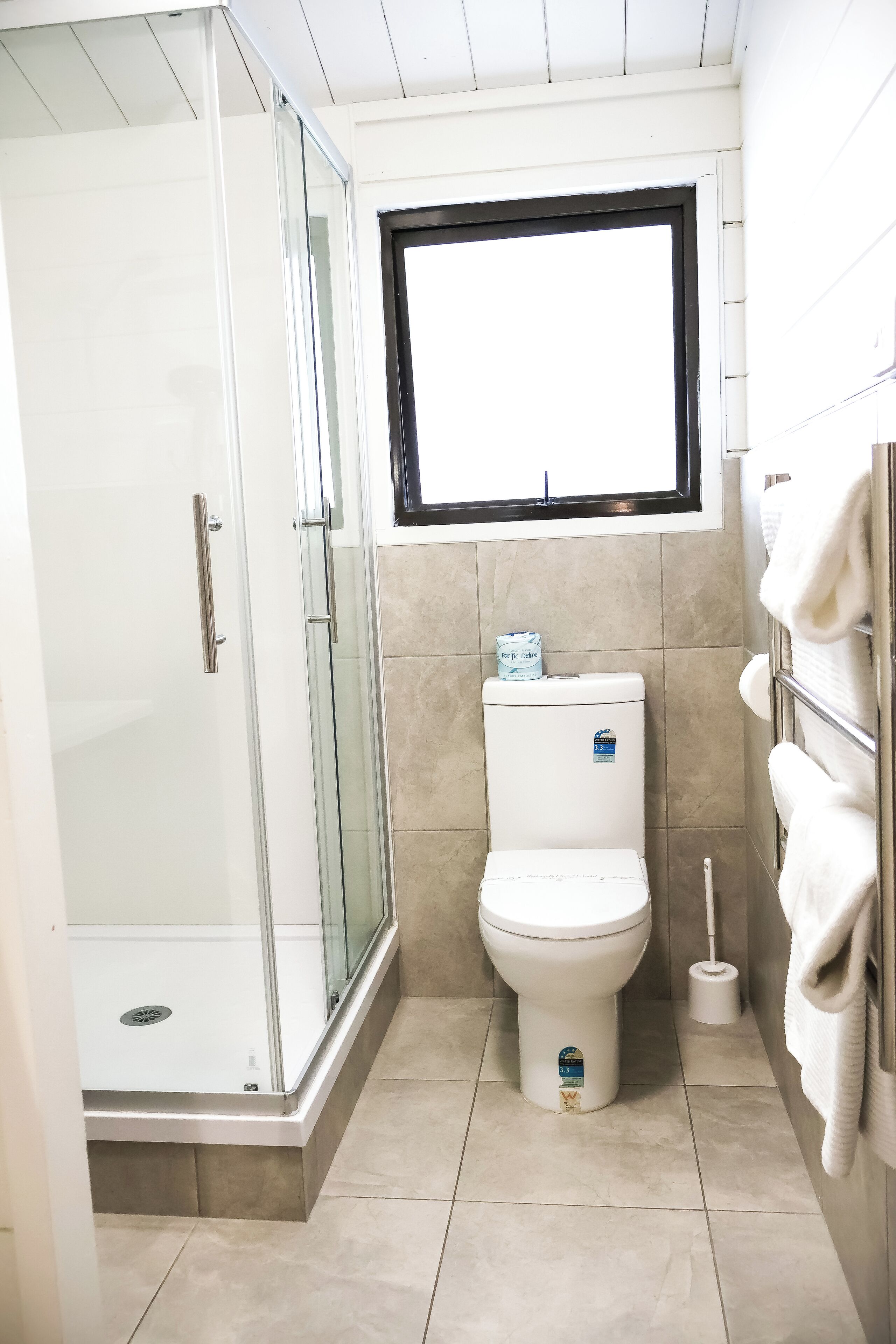 A modern bathroom with a glass shower enclosure, a toilet, and a window above the toilet.