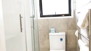 A modern bathroom with a glass shower enclosure, a toilet, and a window above the toilet.