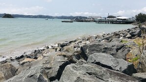A rocky shoreline with a pier extending into calm waters, a building with a white roof, and distant hills. - Bounty Motel (Paihia)