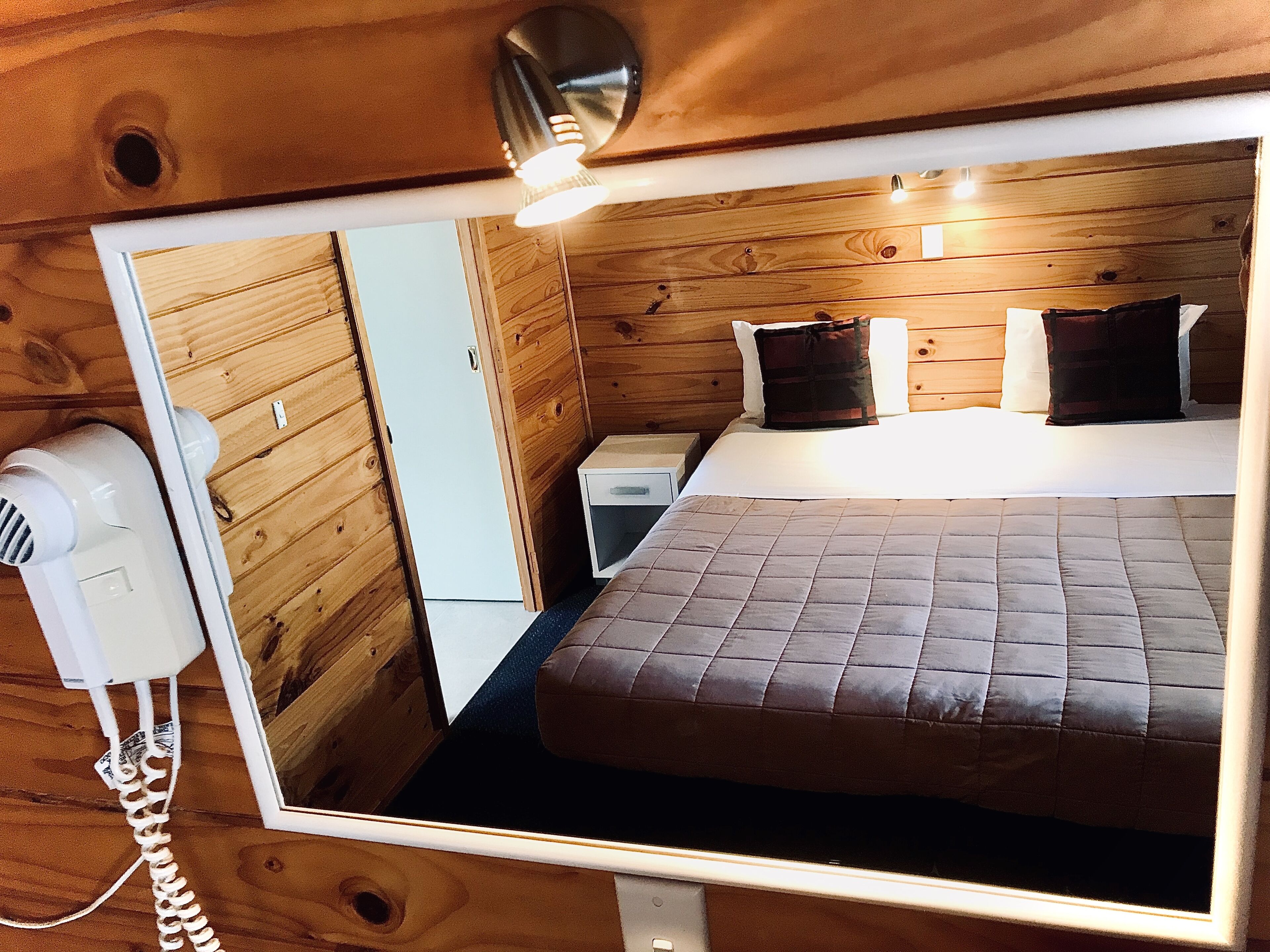 Une chambre dans un chalet en bois, comprenant un lit, une table de chevet, un sèche-cheveux et un miroir qui reflète la pièce.