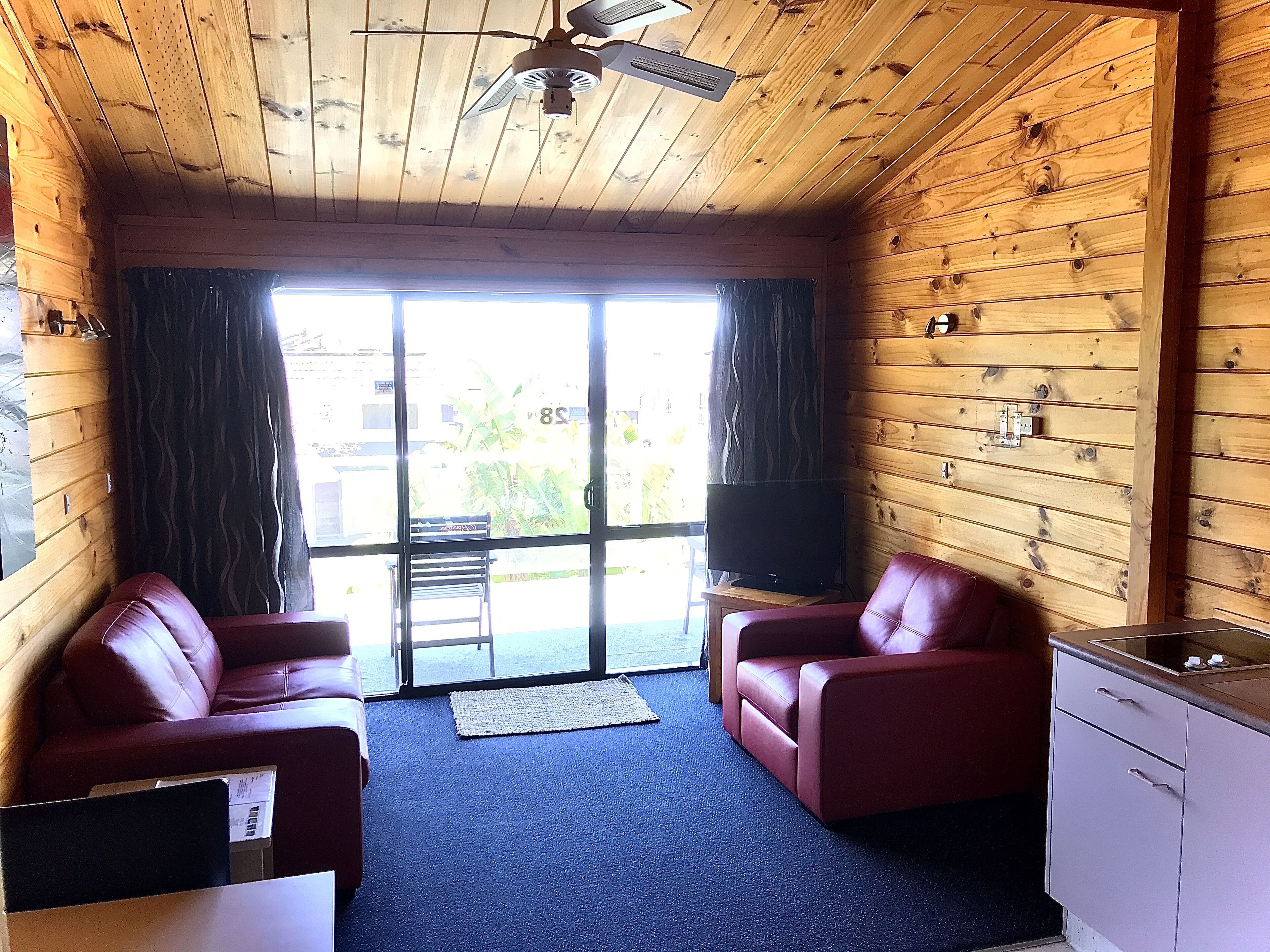 A room with wooden walls, a ceiling fan, a television on a stand, two red leather sofas, a small kitchenette, and a large window with curtains.