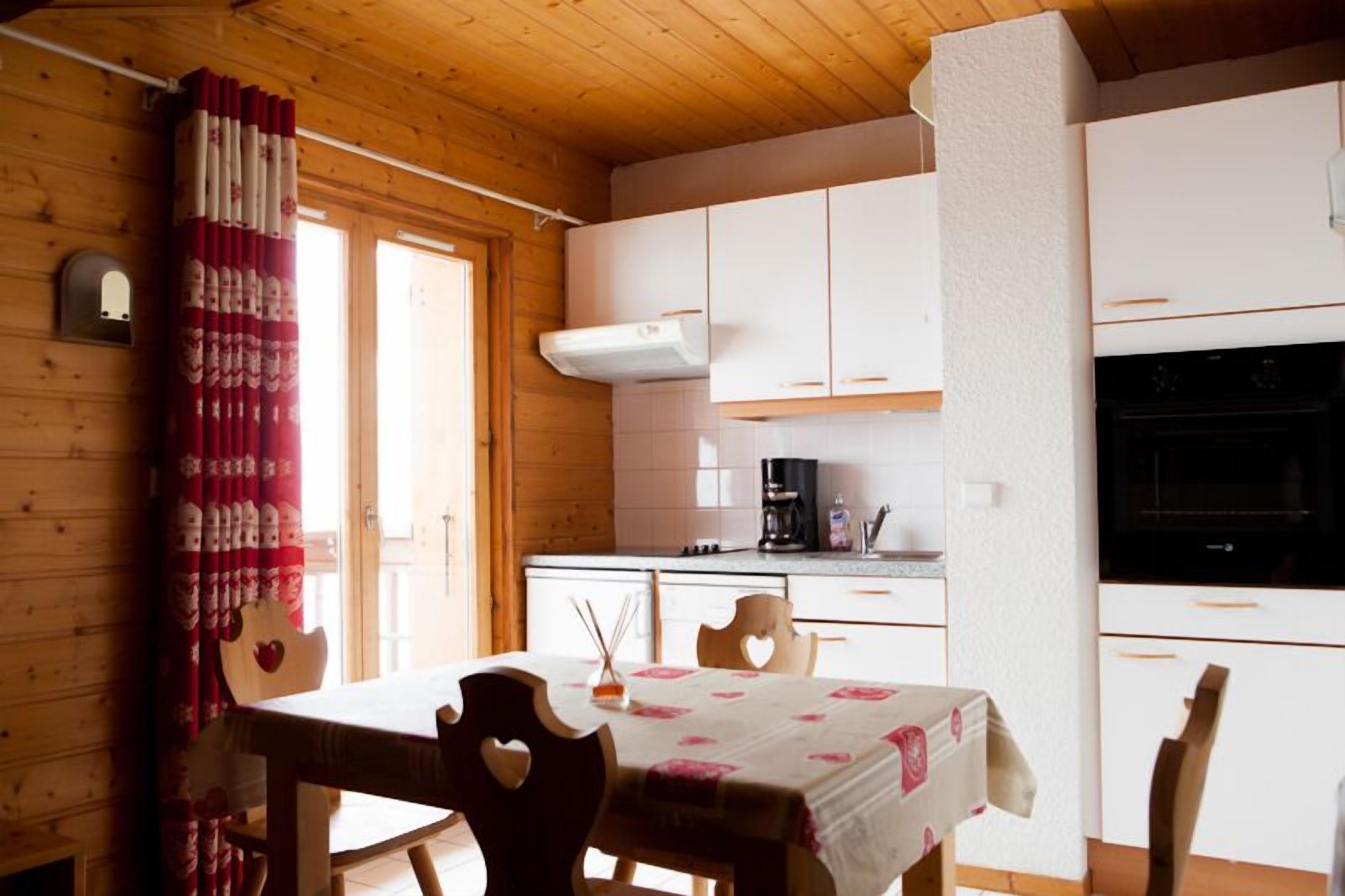 Une cuisine chaleureuse avec des meubles en bois, une table à manger entourée de chaises en forme de cœur et une fenêtre agrémentée de rideaux.