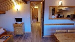 A room with a wooden desk, a television on a wooden cabinet, and a kitchen area with a microwave and kettle. - Le Floralp (Combloux)