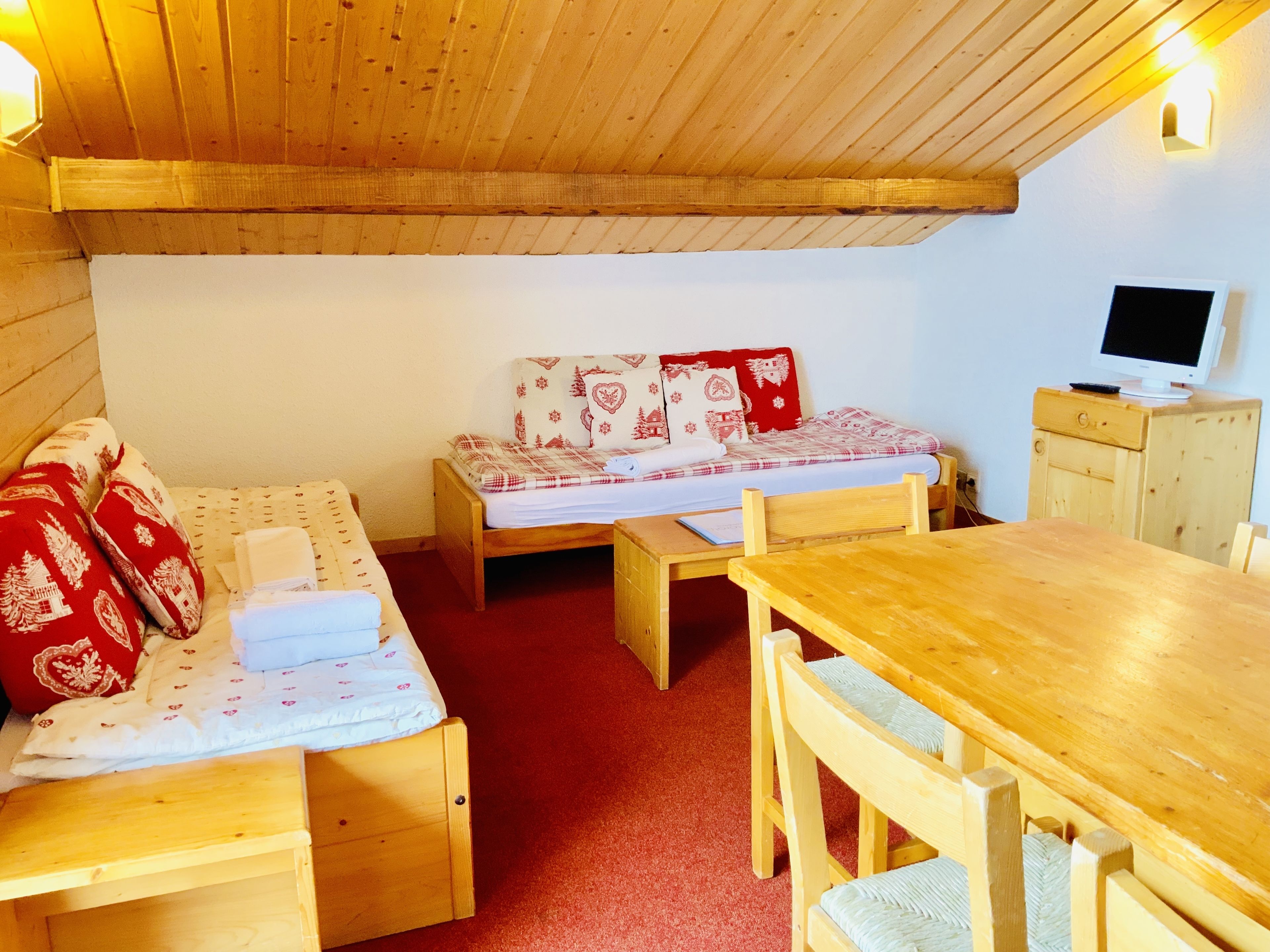 Una habitación acogedora con techo de madera, un sofá con cojines estampados, una cama pequeña, una mesa de madera y un televisor sobre un mueble.