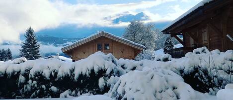 A wooden chalet with a snow-covered roof, surrounded by snow-laden trees and a mountainous backdrop.
