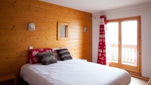 A bedroom with a wooden wall, a bed with white bedding, two decorative pillows, a window with a red curtain, and a small wall-mounted light fixture. - Le Floralp (Combloux)