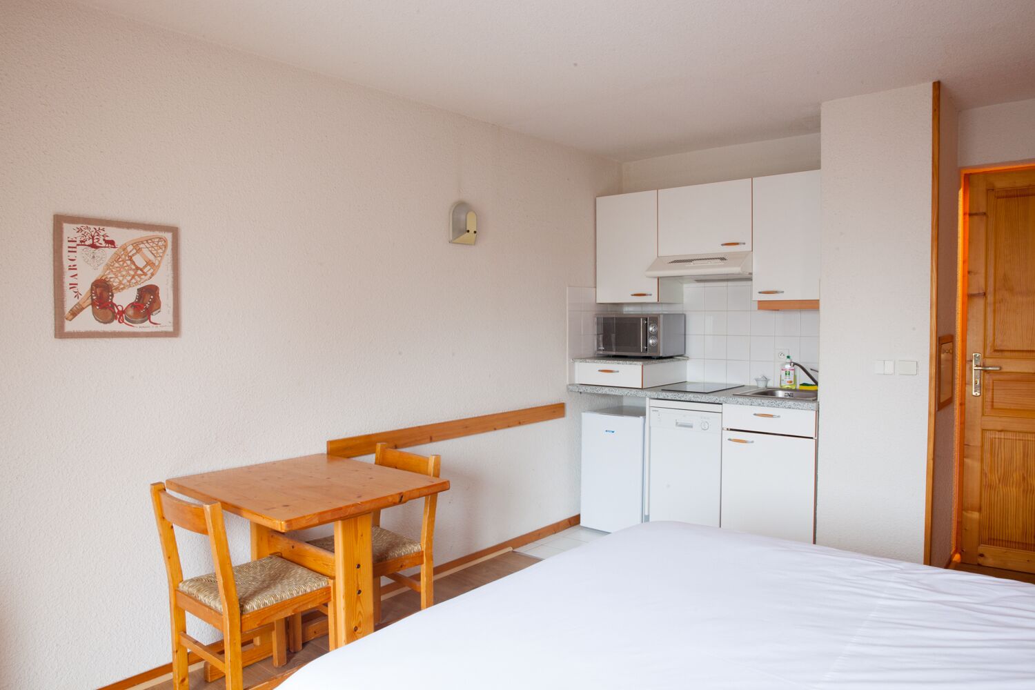 Een compacte kamer met een bed, houten tafel en stoelen, een kitchenette met een magnetron, broodrooster en kleine koelkast, en een houten deur.