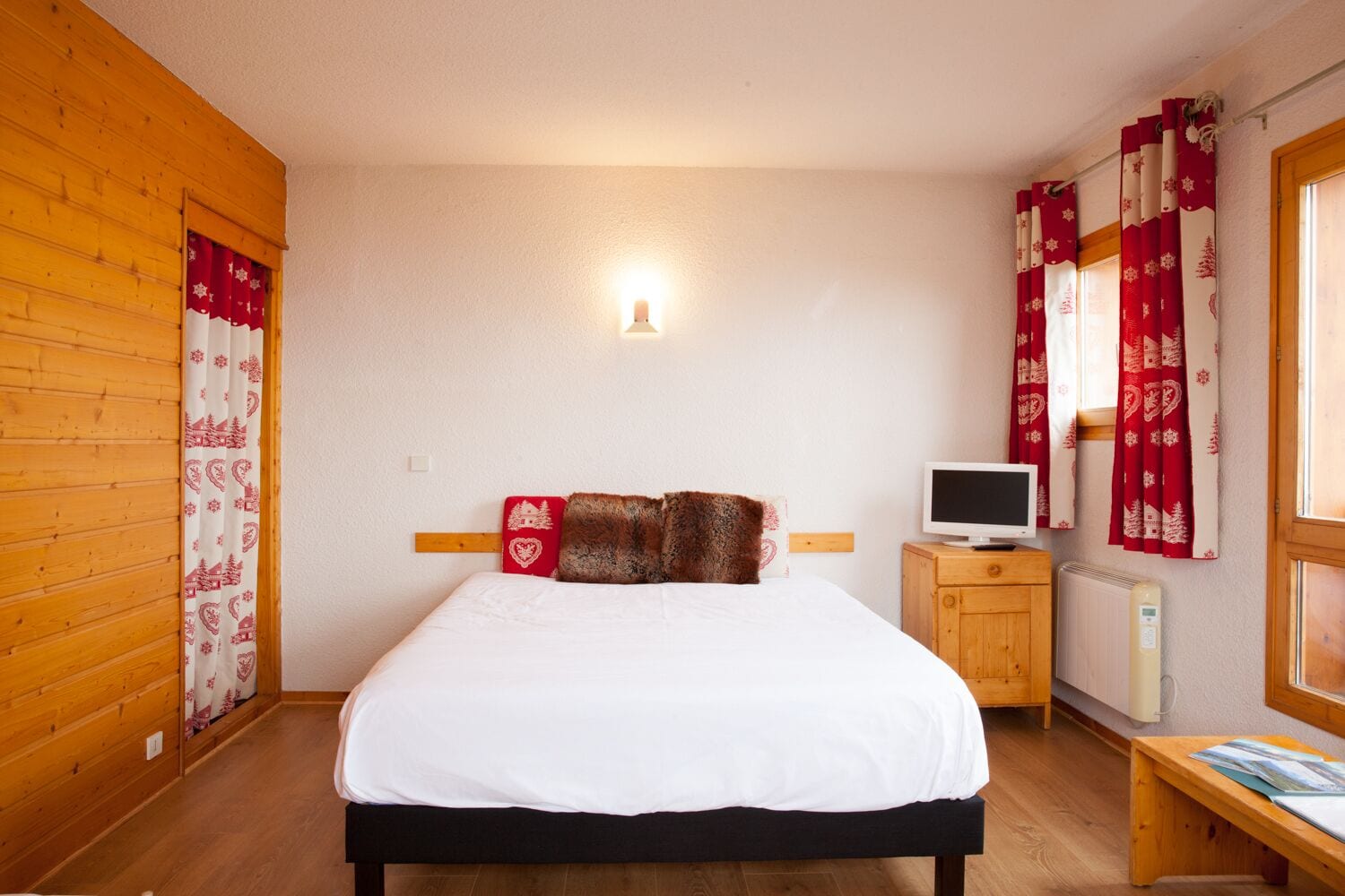 Een slaapkamer met een bed, een nachtkastje met een televisie, een verwarming en houten wanden.