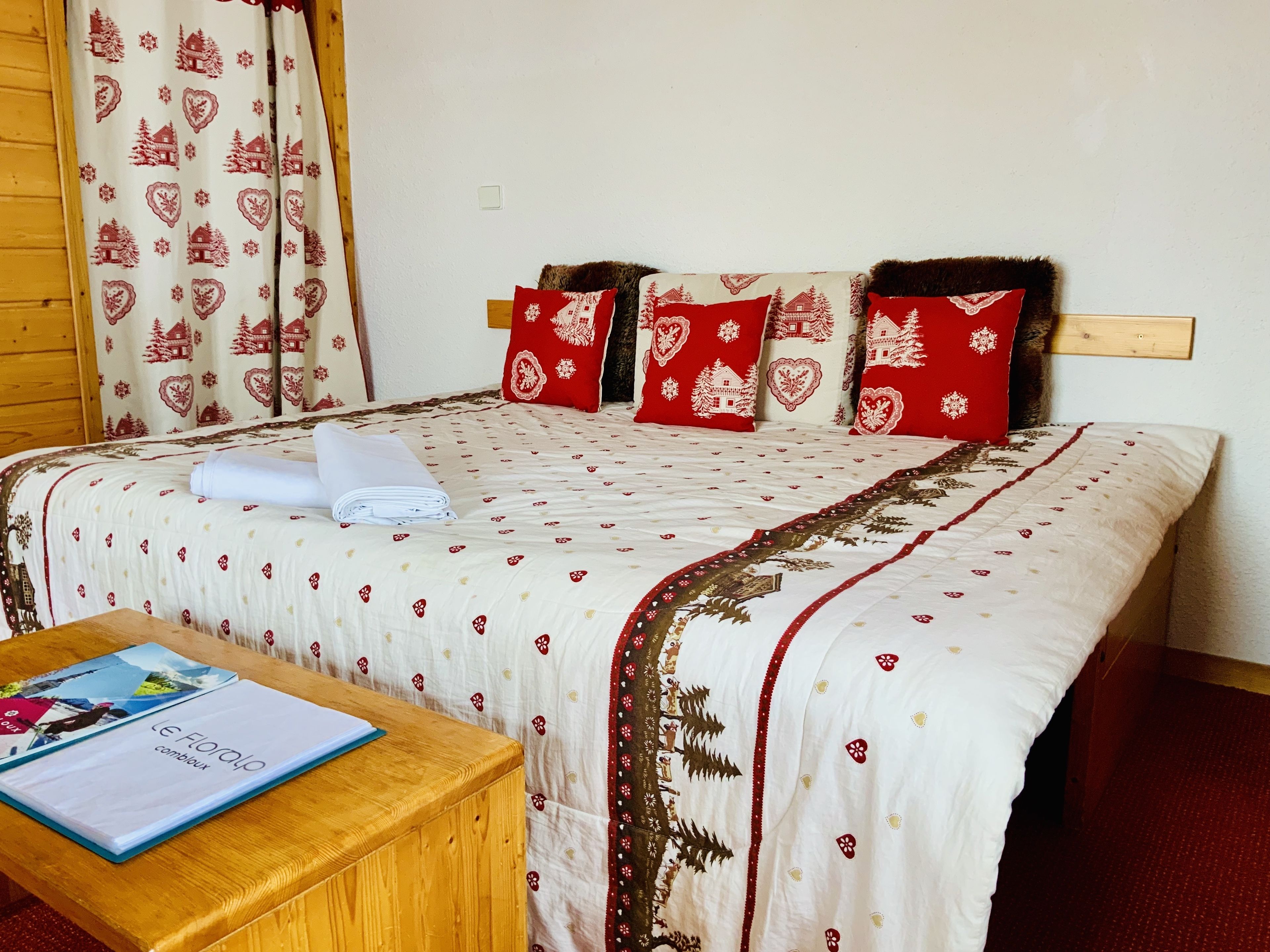 A neatly made bed with a patterned bedspread, red pillows, and a folded white towel on a wooden bedside table.