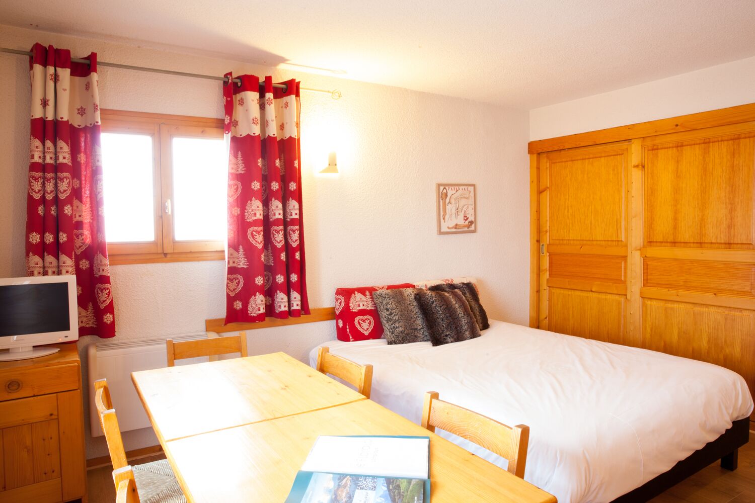 A bedroom with a bed, wooden headboard, a desk with a chair, a television, and red curtains with a pattern.