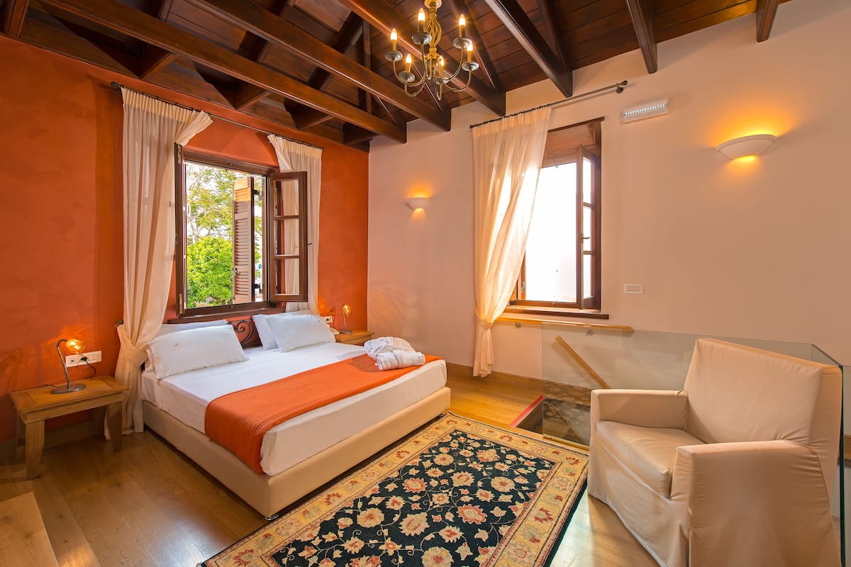 Une chambre à coucher comprenant un lit, une chaise, une table de chevet, un lustre et une fenêtre avec des rideaux.