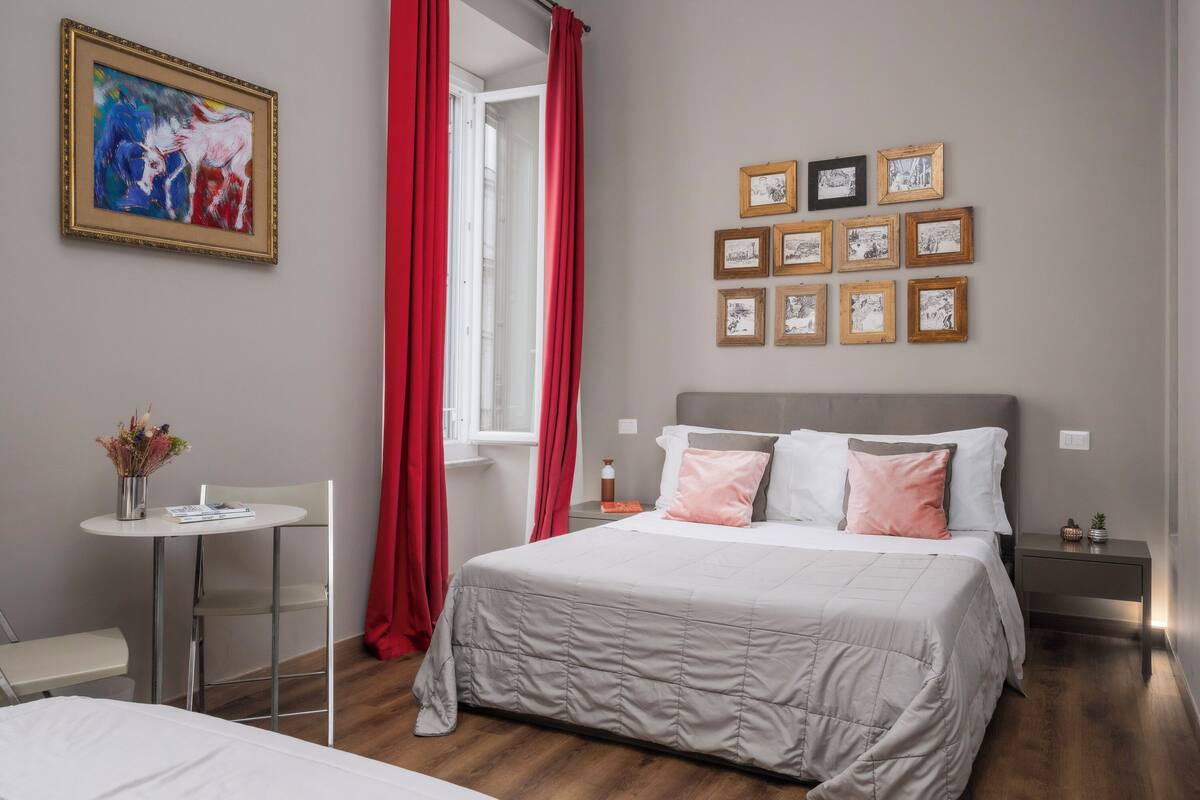 A bedroom with a bed, a bedside table, a chair, a window with red curtains, and a wall with framed pictures.