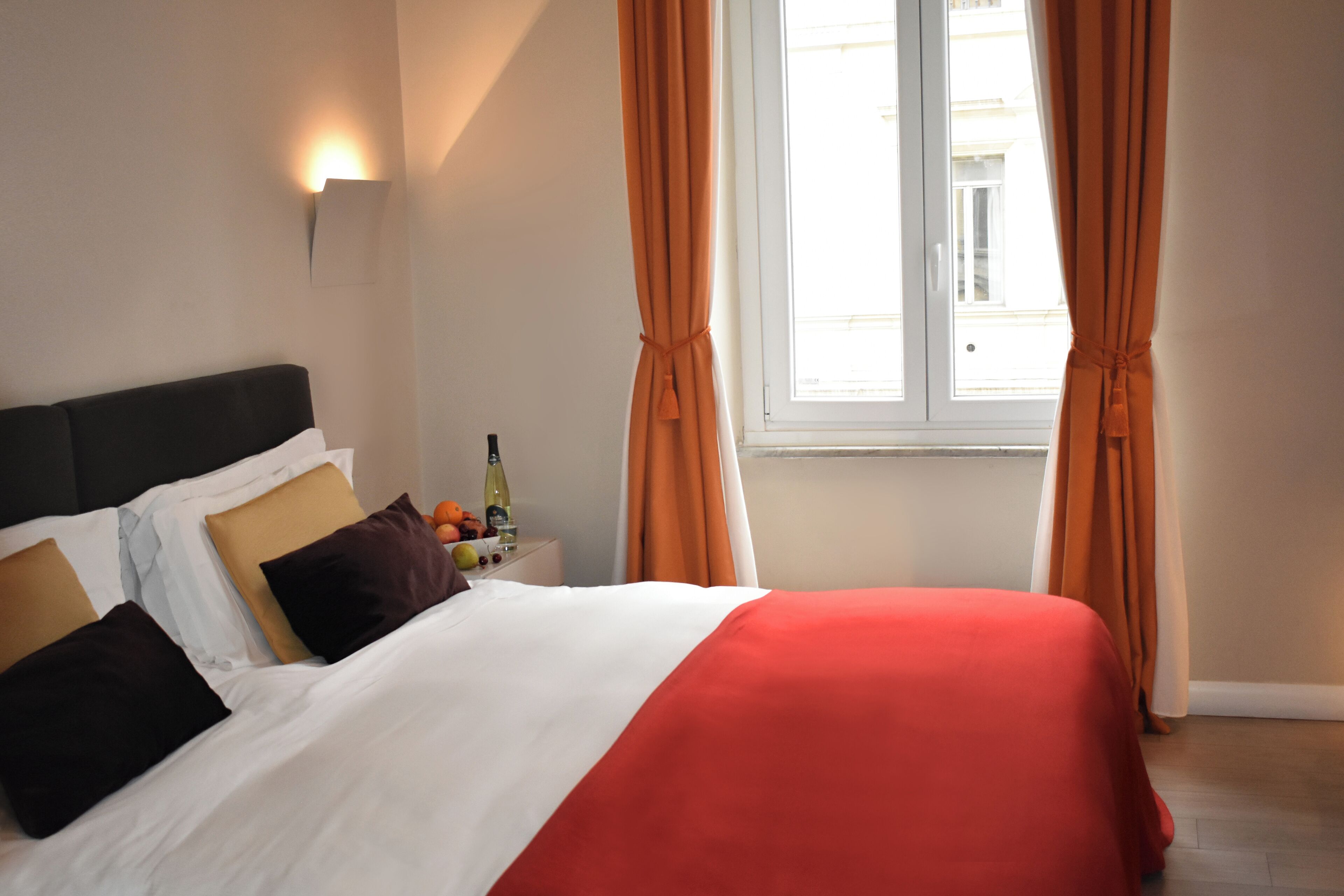 A hotel room with a bed, a nightstand, a bottle of wine, and orange curtains.