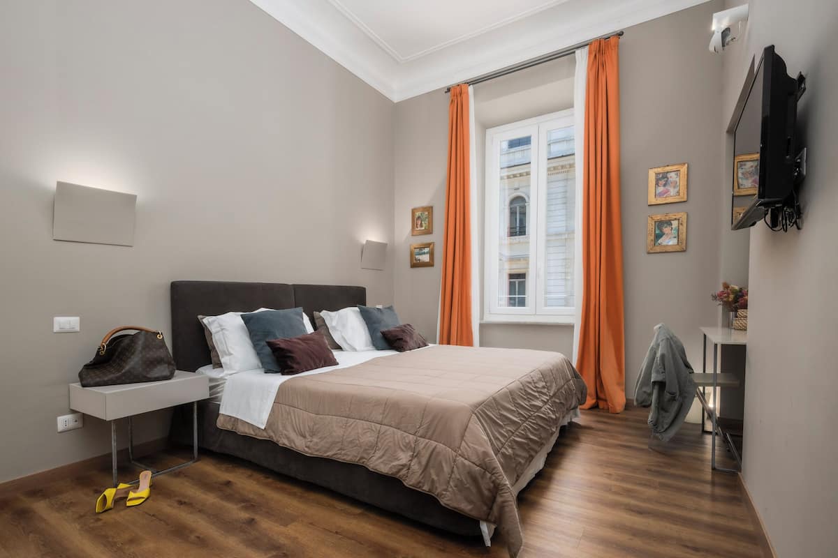 A bedroom with a bed, a bedside table, a TV, and a window with curtains.