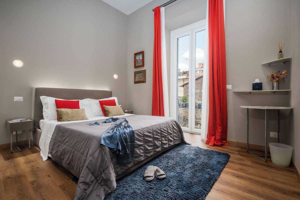 A bedroom with a bed, a nightstand, a desk, a chair, a window with red curtains, and a view of the outside.