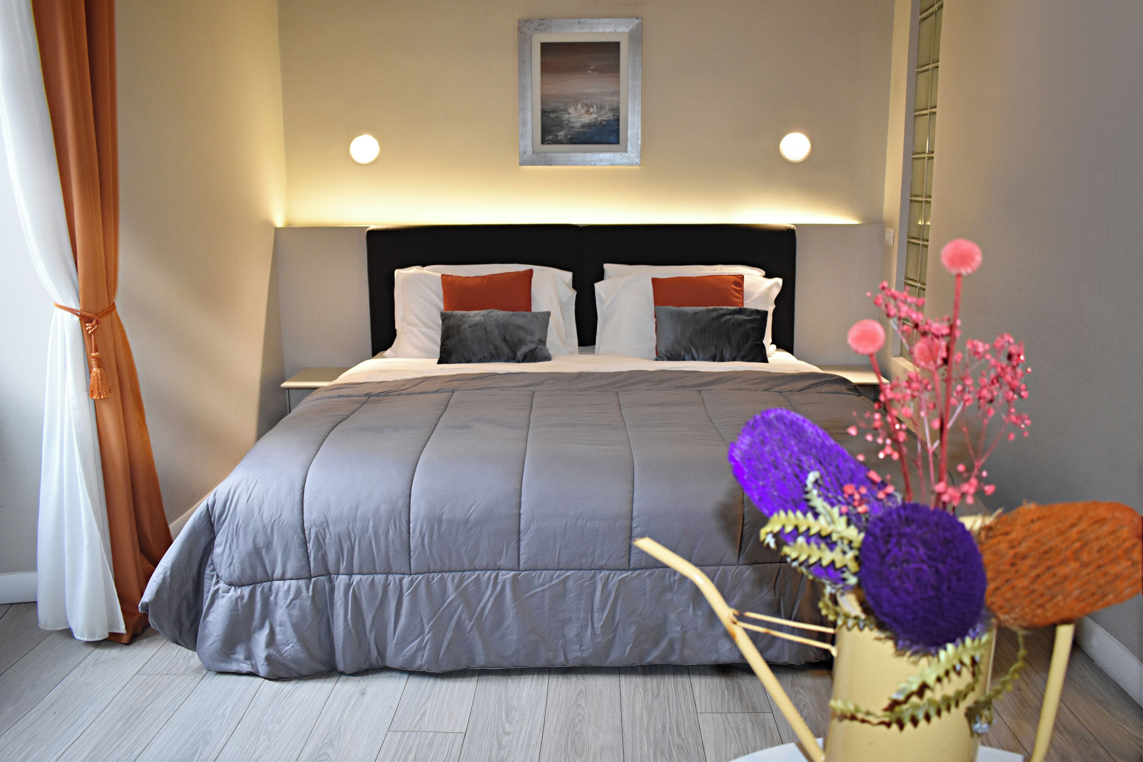 A bedroom with a large bed, two pillows, a nightstand, a wall-mounted painting, and a vase with flowers.