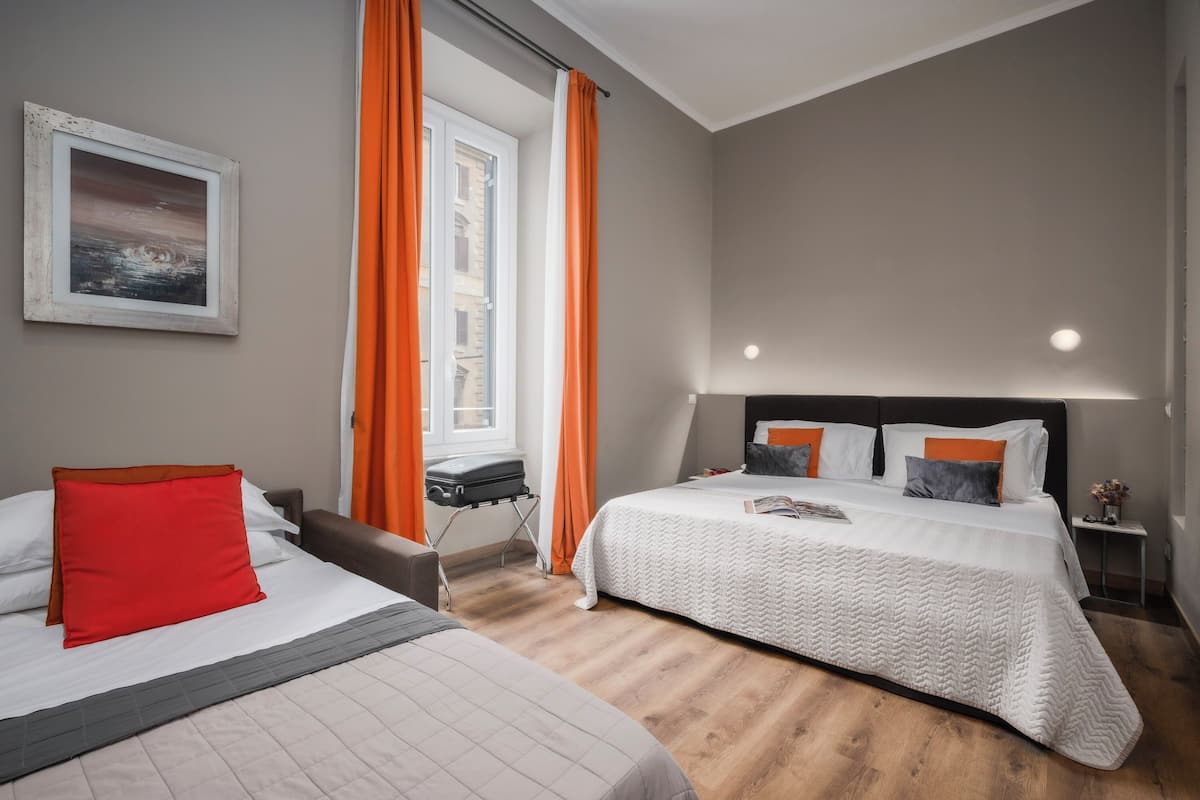 A hotel room with two beds, a window with orange curtains, a painting on the wall, and a nightstand with a lamp.