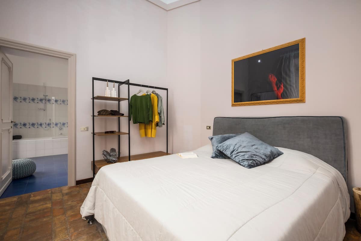 A bedroom with a bed, a grey headboard, a blue pillow, a wooden floor, a white wall, a framed picture, a clothing rack, and a bathroom with a shower and a bathtub.