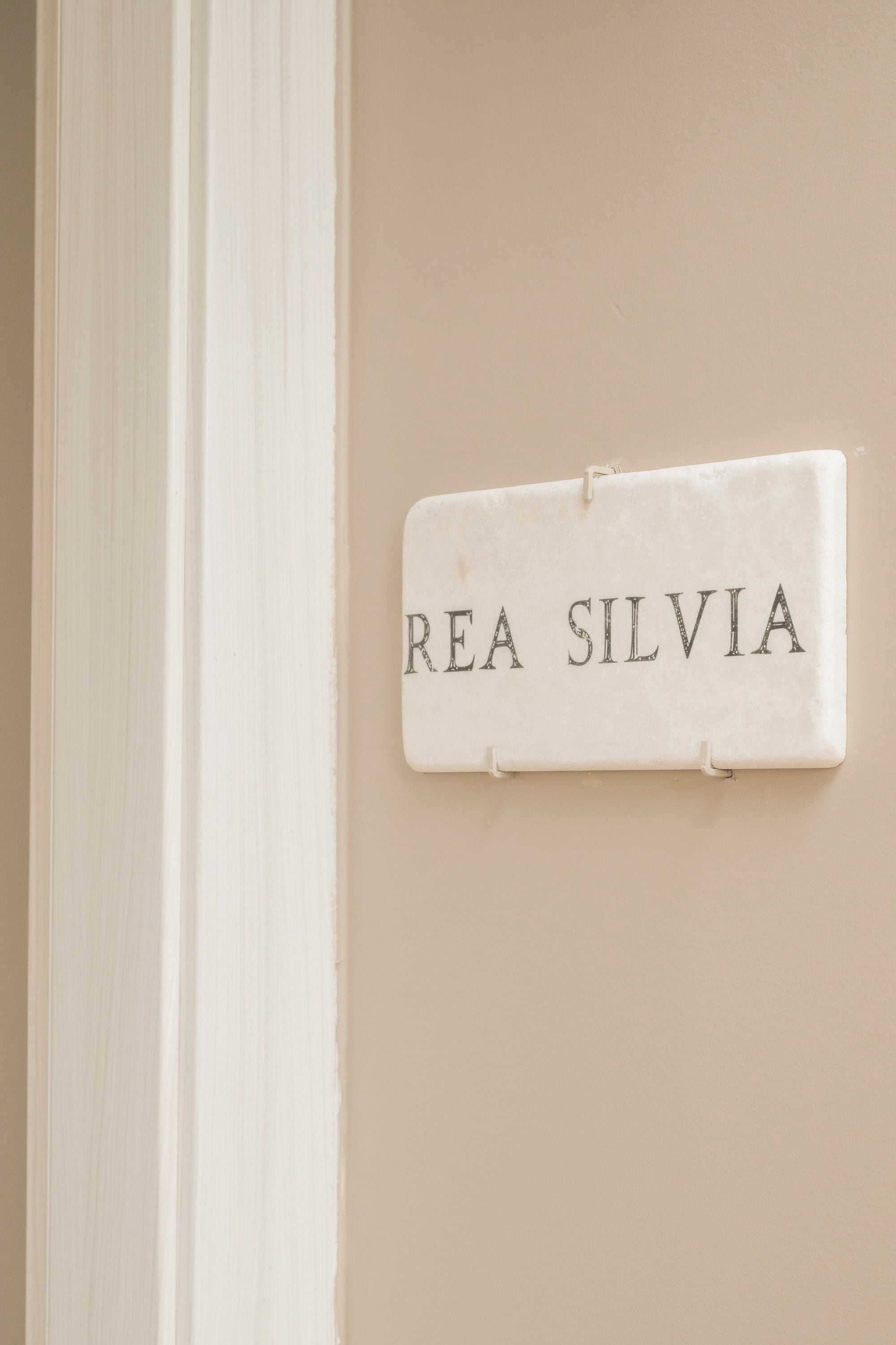 En vit skylt med texten ”REA SILVIA” som är fastsatt på en beige vägg.