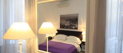 A hotel room with a bed, two bedside lamps, a nightstand, a mirror, and a cityscape painting on the wall.