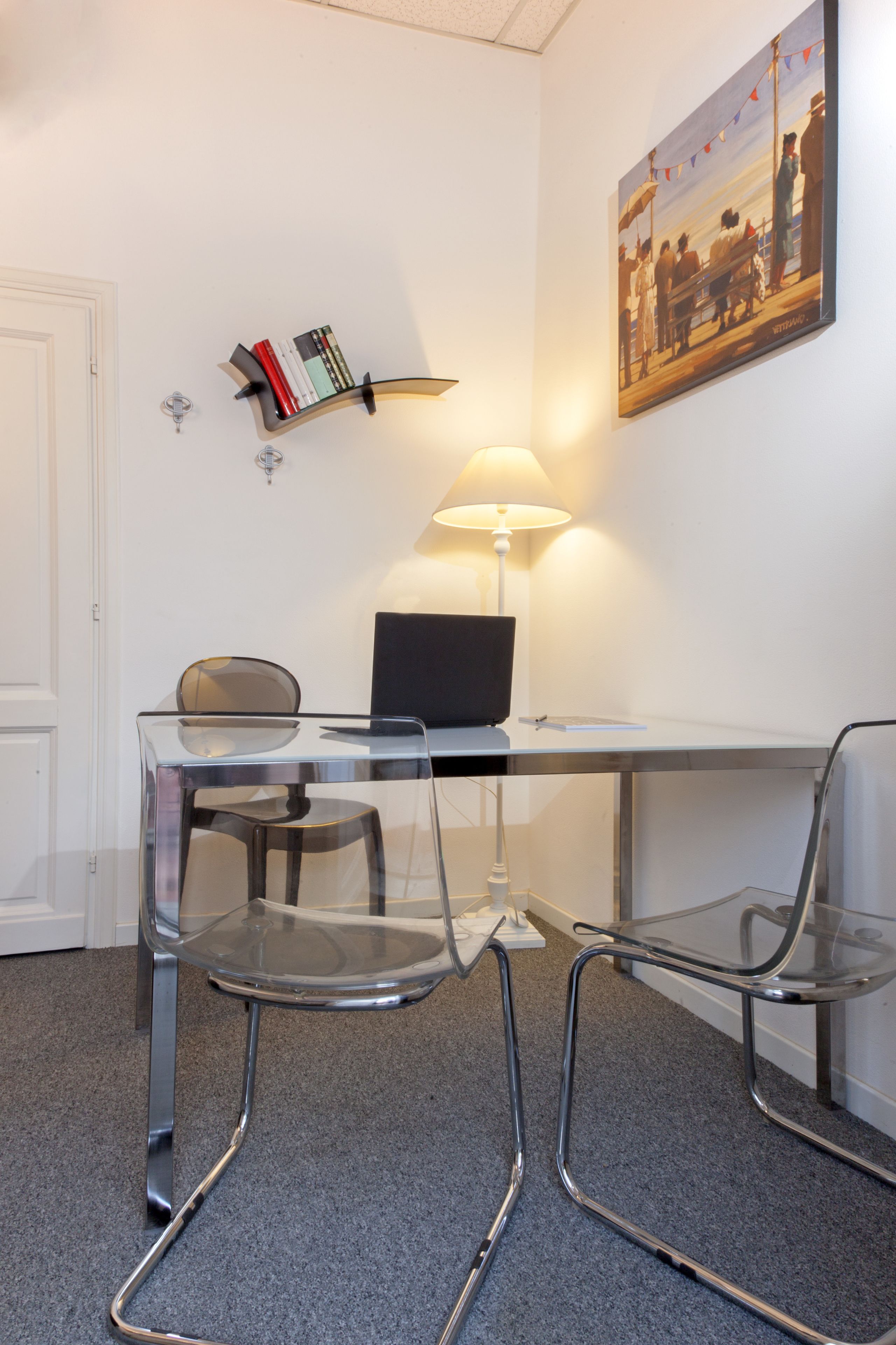 A modern office space with a desk, two chairs, a laptop, a lamp, a bookshelf, and a painting on the wall.