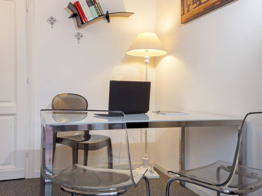A modern office space with a desk, two chairs, a laptop, a lamp, a bookshelf, and a painting on the wall.