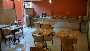A dining area with multiple tables and chairs, a kitchen in the background, and a tiled floor. - Pousada Praia Bella (Búzios)
