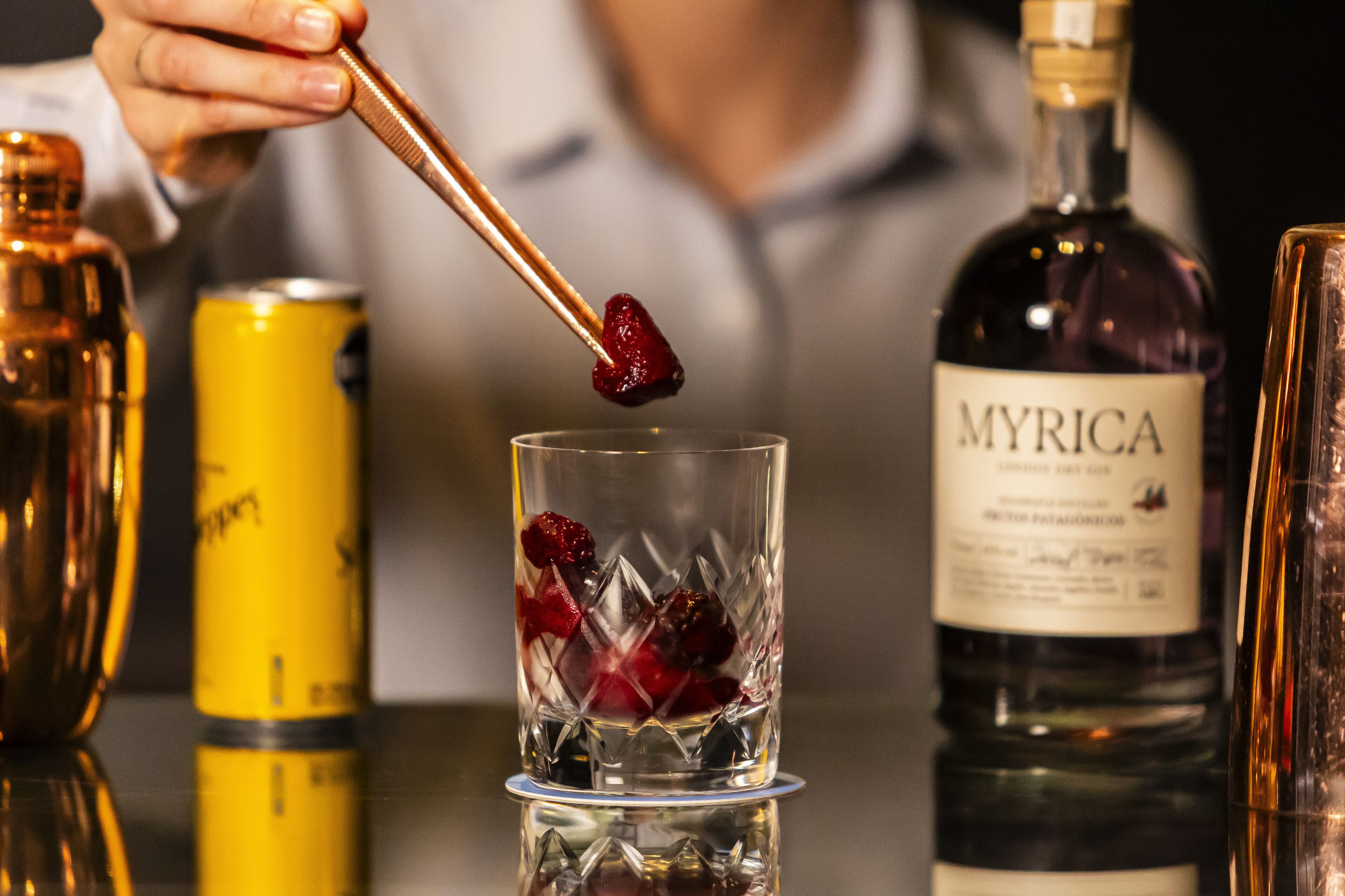 Een cocktail wordt bereid, met de hand van een bartender, ijsblokjes, een blikje en een fles met de tekst 'MYRICA' op de achtergrond.
