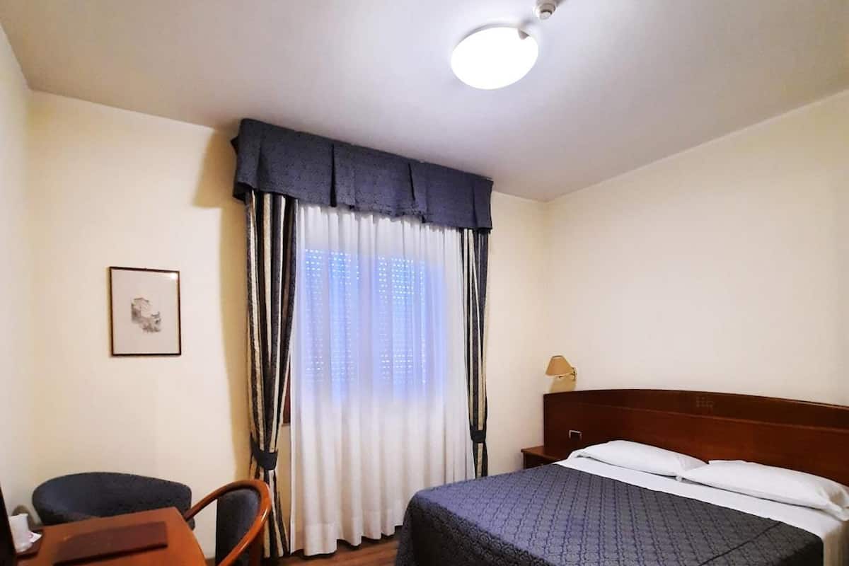 A hotel room with a wooden floor, a bed with a dark patterned bedspread, a desk with a chair, a bedside table with a lamp, and a window with sheer curtains.