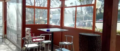 A covered outdoor seating area with a table and chairs, large windows, and a view of the outside.