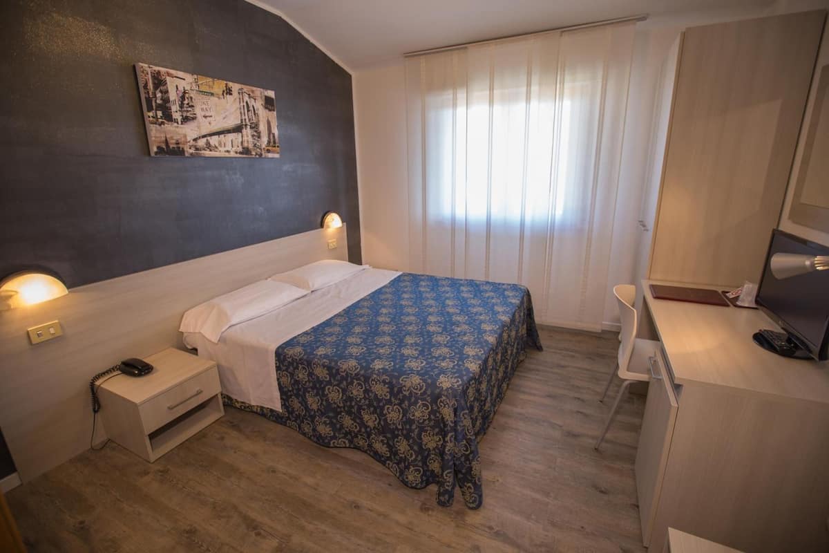 A hotel room with a bed, bedside table, lamp, desk with a chair, and a TV.