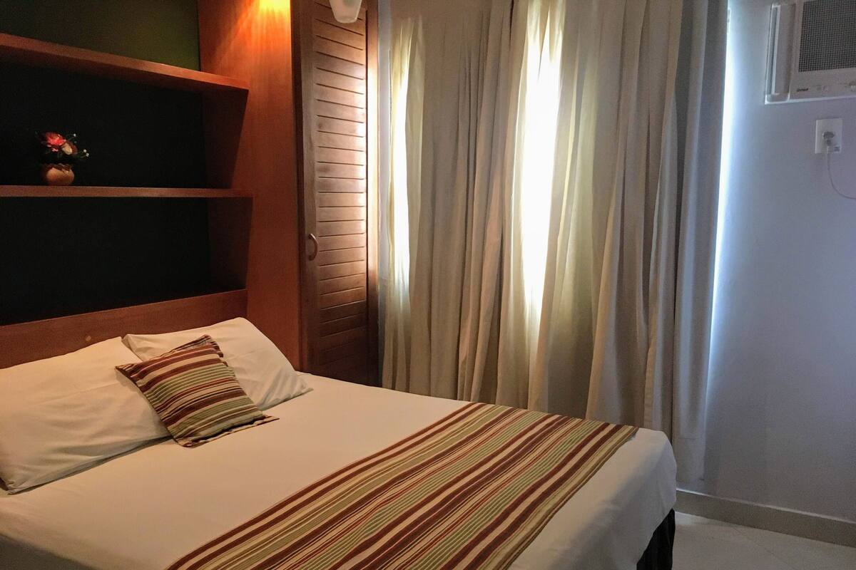 Ein Hotelzimmer mit einem Bett, einem Nachttisch, einem Regal mit einer Vase und einem Fenster mit Vorhängen.