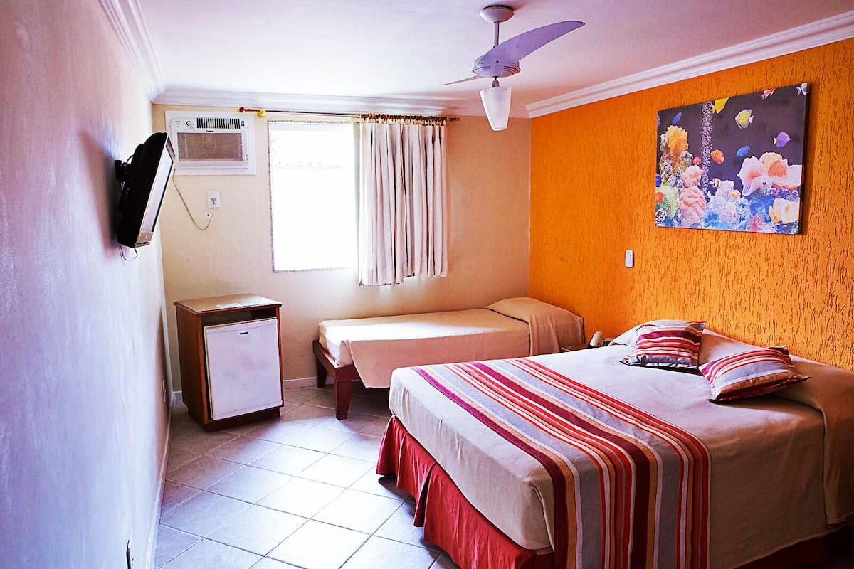 Ein Hotelzimmer mit Bett, Schreibtisch, kleinem Kühlschrank, Deckenventilator und einem Fenster mit Vorhängen.