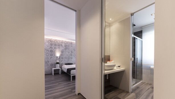 Ein modernes Hotelzimmer mit Holzboden, einem Bett mit gemustertem Kopfteil, einem Nachttisch mit Lampe und einem Badezimmer mit Duschkabine.