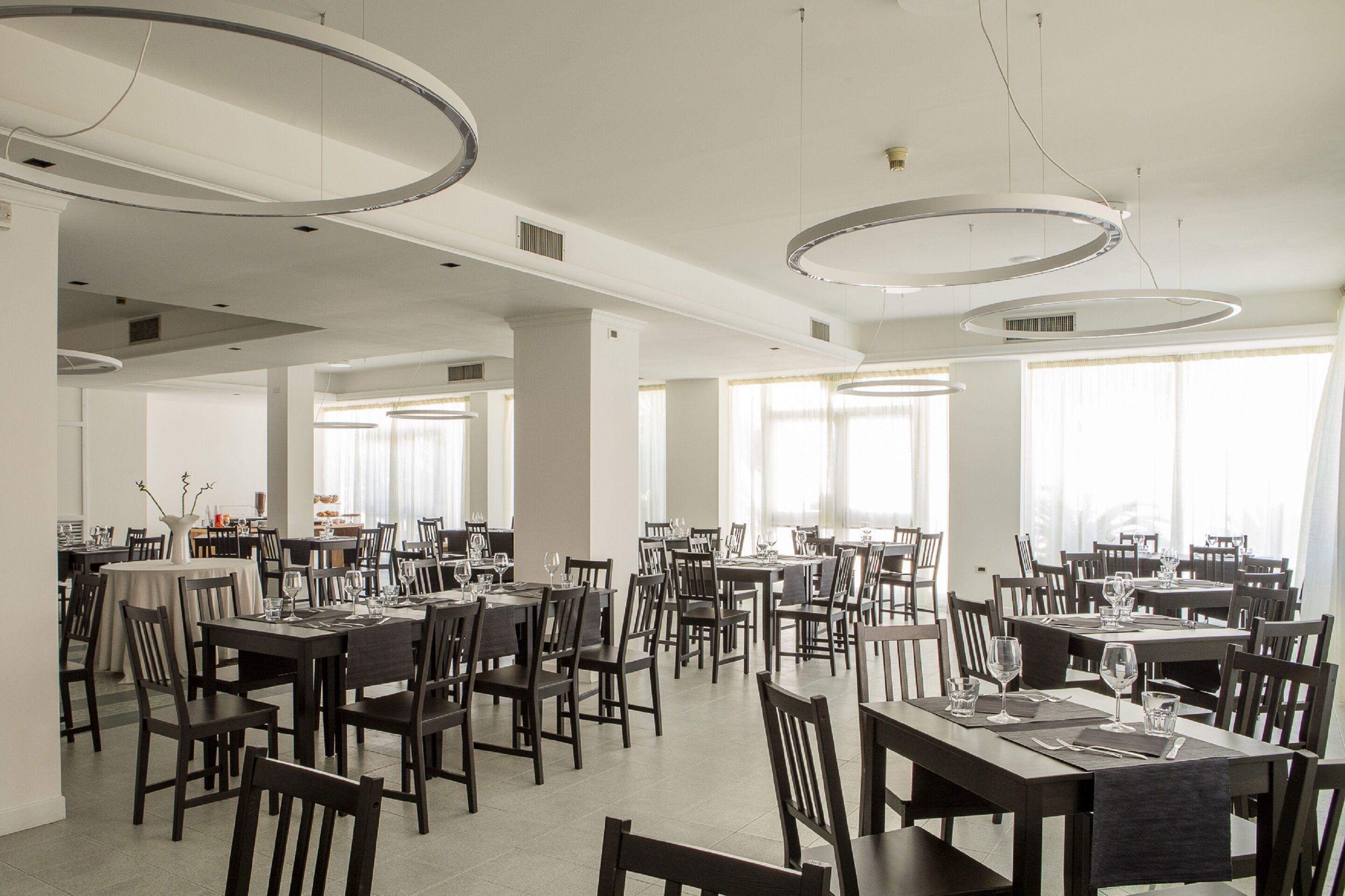 Un vaste espace dédié à la salle à manger, avec de nombreuses tables et chaises, de grandes fenêtres et des luminaires circulaires modernes au plafond.