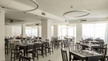 Un vaste espace dédié à la salle à manger, avec de nombreuses tables et chaises, de grandes fenêtres et des luminaires circulaires modernes au plafond.