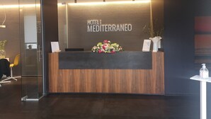Ein Hotelrezeptionstresen mit Holzvertäfelung, einer Blumenkomposition und einem Schild mit der Aufschrift 'HOTEL: MEDITERRANEO'.