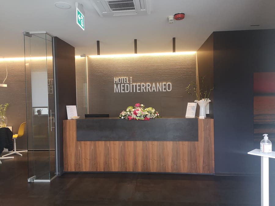 Ein Hotelrezeptionstresen mit Holzvertäfelung, einer Blumenkomposition und einem Schild mit der Aufschrift 'HOTEL: MEDITERRANEO'.