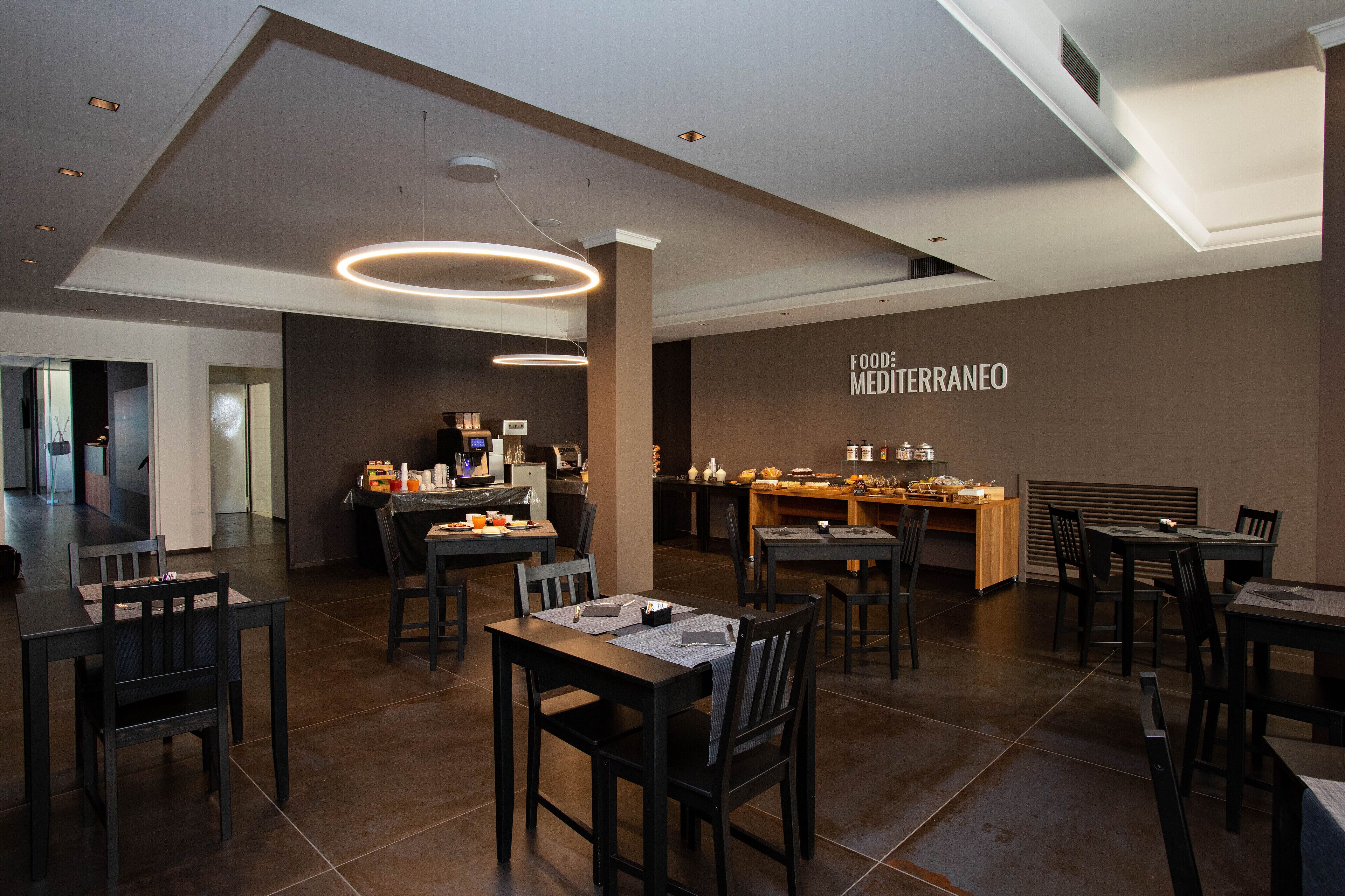 Een modern restaurantinterieur met meerdere eettafels en stoelen, een keukenhoek en een muur met de tekst 'FOOD: MEDI TERRANEO'.