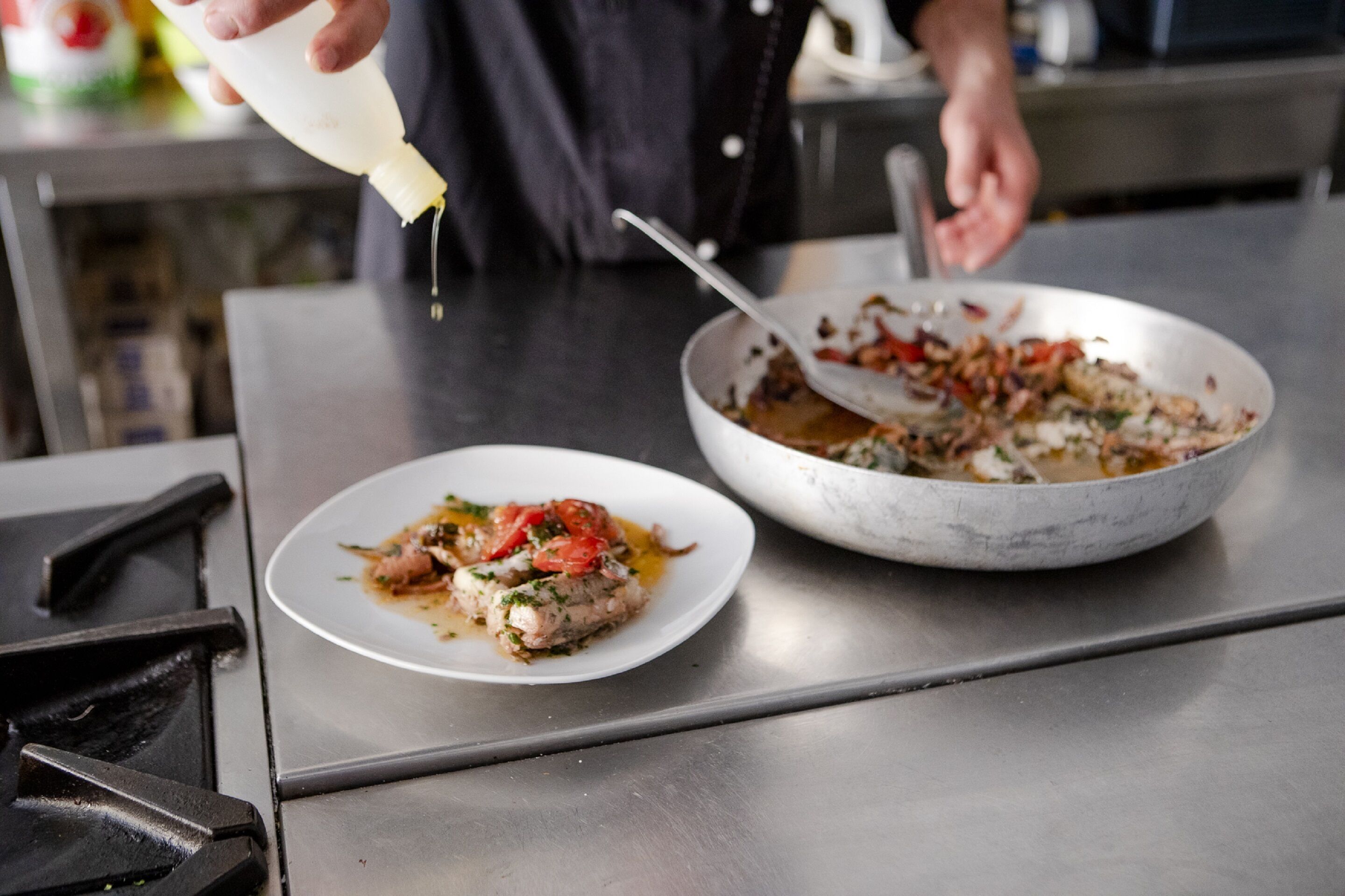A person pouring sauce onto a dish with food and a pan on a stainless steel counter.