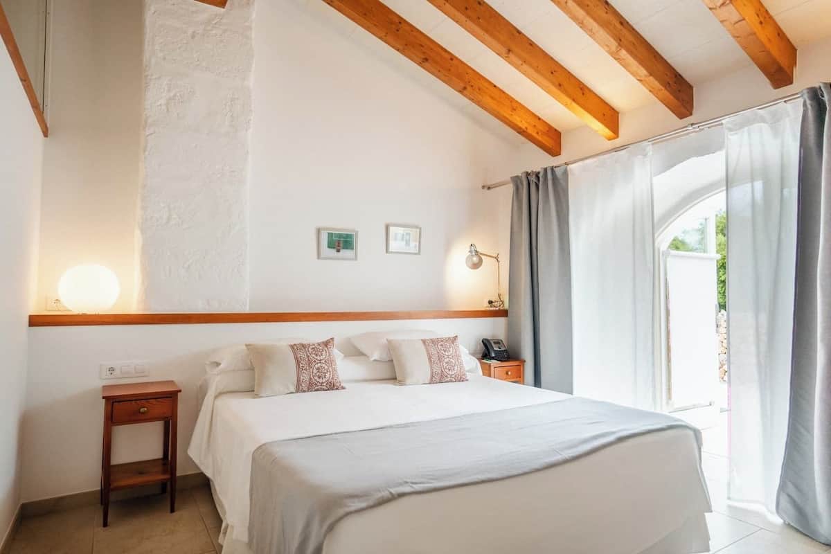 A bedroom with a wooden ceiling, a bed with white bedding, a bedside table with a lamp, and a window with curtains.