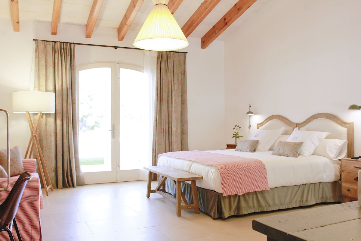 A bedroom with a bed, bench, bedside table, lamp, and curtains.