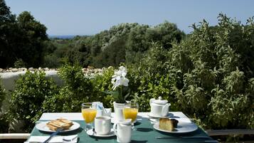 A table set with breakfast items including bread, juice, and a pastry, overlooking a scenic coastal area with dense greenery and trees.