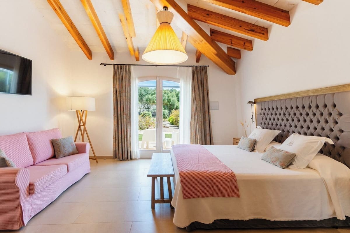 A bedroom with a bed, a sofa, a lamp, and a view of the outdoors.