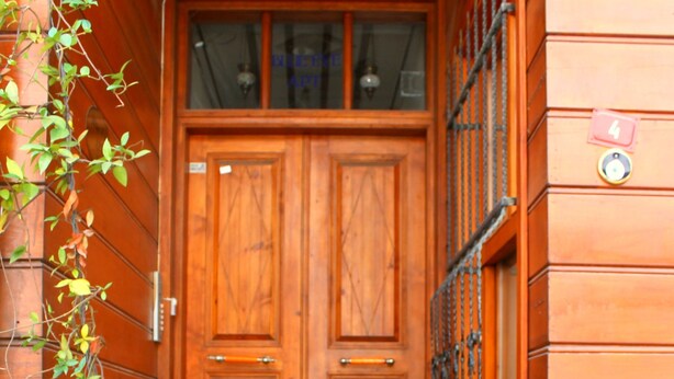 Una puerta doble de madera con un felpudo verde en la parte inferior, entre paredes de ladrillo y macetas con plantas.
