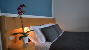 Ein ordentlich bezogenes Bett mit dunkler Decke, einem weißen und einem grauen Kissen auf einem hölzernen Kopfteil. Ein weißer Nachttisch mit einer Topforchidee ist sichtbar.